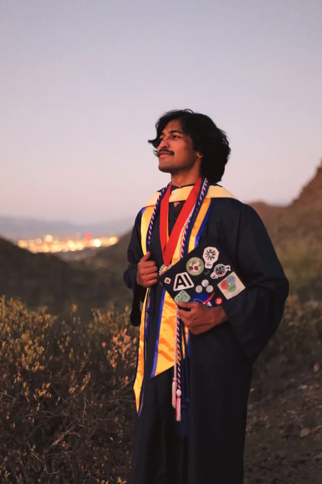 Akash in graduation regalia at dusk on a desert hillside, with Tucson's city lights in the distance